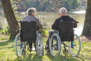 Elderly couple, each in a wheelchair, looking at a lake